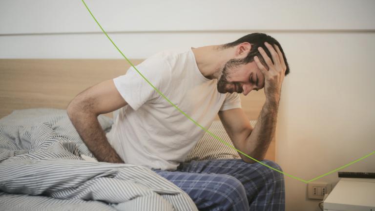 Man met wit t-shirt en geruite broek zit op rand van het bed met een hand op zijn rug en een hand op zijn hoofd