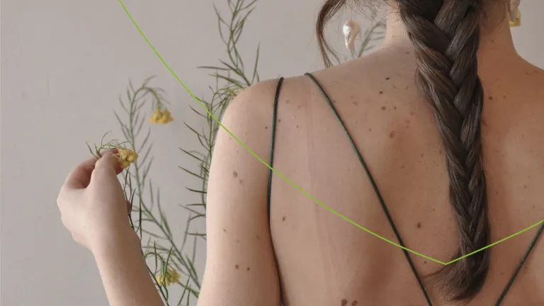 rug van een vrouw met moedervlekken met op de achtergrond een roze muur en bloemen die ze aanraakt