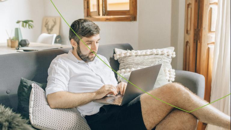 man in korte broek thuis op de bank met laptop op schoot en benen over elkaar