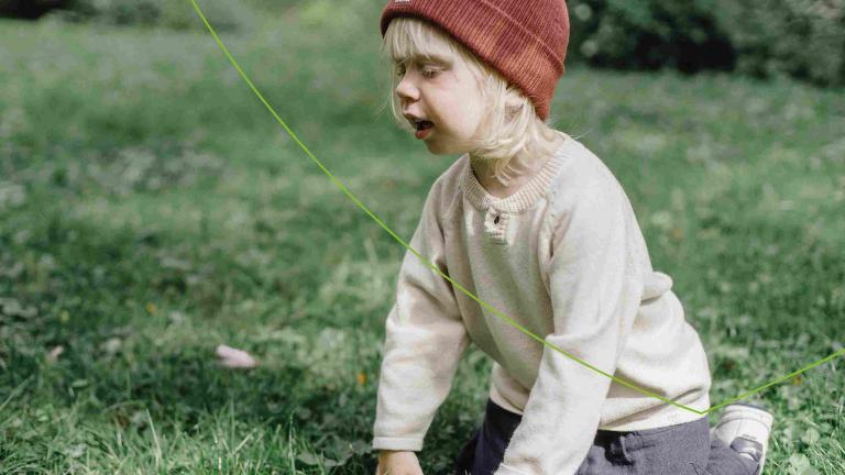 Kindje met blond haar in beige trui en rode muts ziet op zijn knieën in het gras en niest 